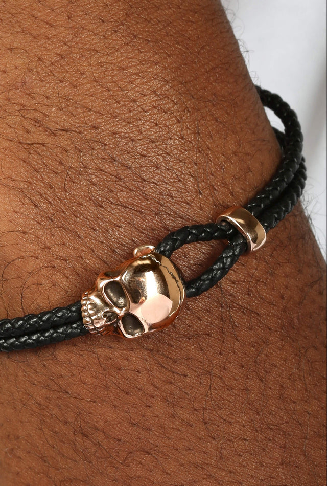Black braided bracelet with a gold skull charm on a wrist.
